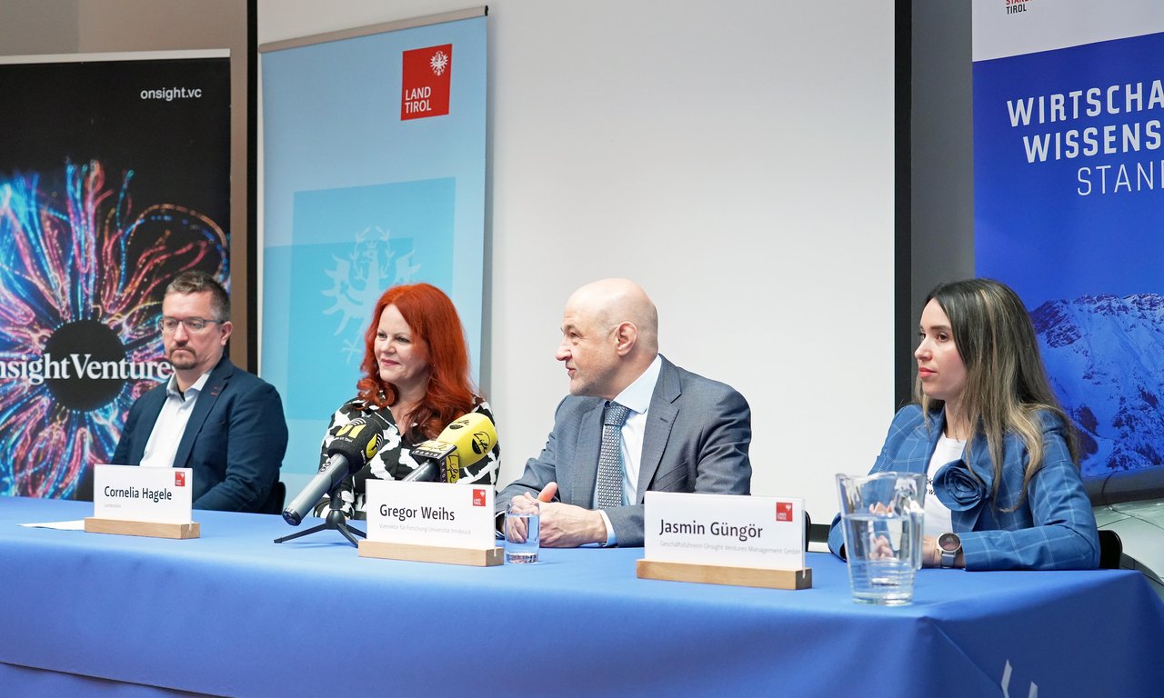 Vier Personen sitzen an einem Tisch mit blauer Tischdecke, vor ihnen stehen Mikrofone und Namensschilder, im Hintergrund Roll-ups von Standortagentur Tirol und Land Tirol
