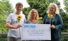Drei Personen stehen jeweils mit einer Sonnenblume vor grünem Hintergrund und halten einen Scheck mit der Spendensumme von 11.561,11 € hoch.