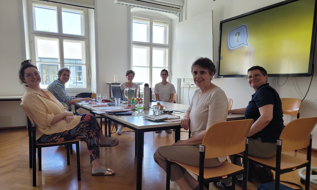 Zu sehen sind die Mitglieder des Fachbereichs Katechetik, Religionspädagogik und -didaktik beim Klausurtag. Sie sitzen an einem großen Tisch und arbeiten gemeinsam. Sie lächeln.
