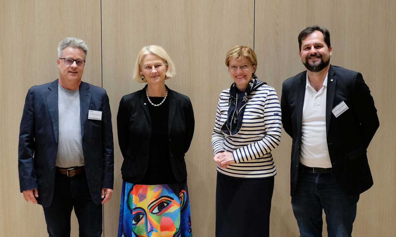 Foto von vier Personen nebeneinander vor einer Holzwand.