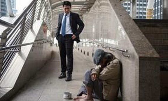 Person mit Anzug steht vor einer am Boden sitzenden Person ohne Schuhe mit einem Becher vor sich.