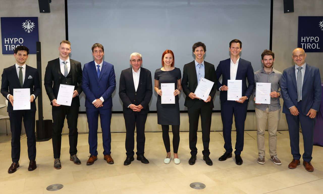 Neun Personen stehen für ein Gruppenfoto nebeneinander, mehrere davon halten Urkunden.