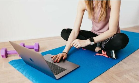 Person sitzt auf einer Yogamatte mit einem Laptop vor sich auf der Matte.