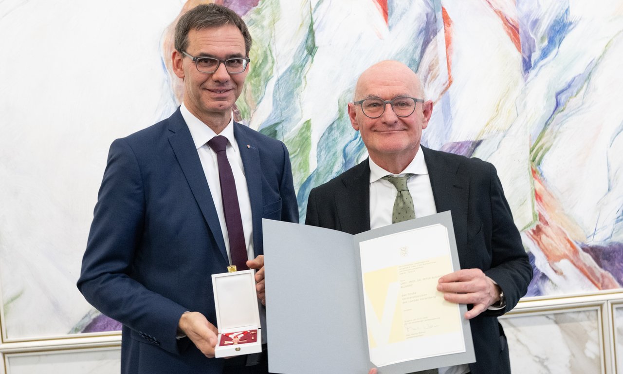 Links steht Landeshauptmann Wallner, der den Orden in der Hand hält, rechts steht Peter Bußjäger und präsentiert die Urkunde.