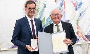 Links steht Landeshauptmann Wallner, der den Orden in der Hand hält, rechts steht Peter Bußjäger und präsentiert die Urkunde.