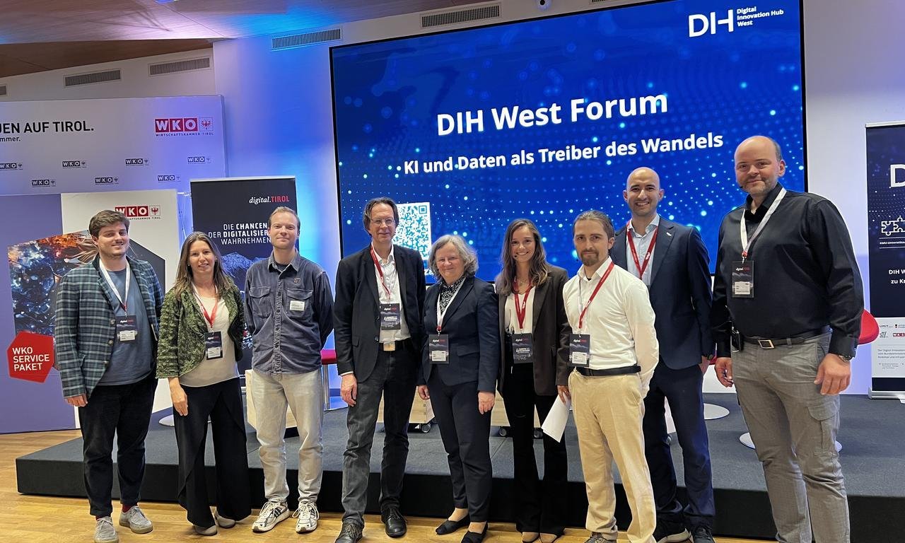 Einige Vortragende des DIH West Forums stehen vor einer Leinwand, die Leinwand ist im CI Blau und Logo vom DIH West