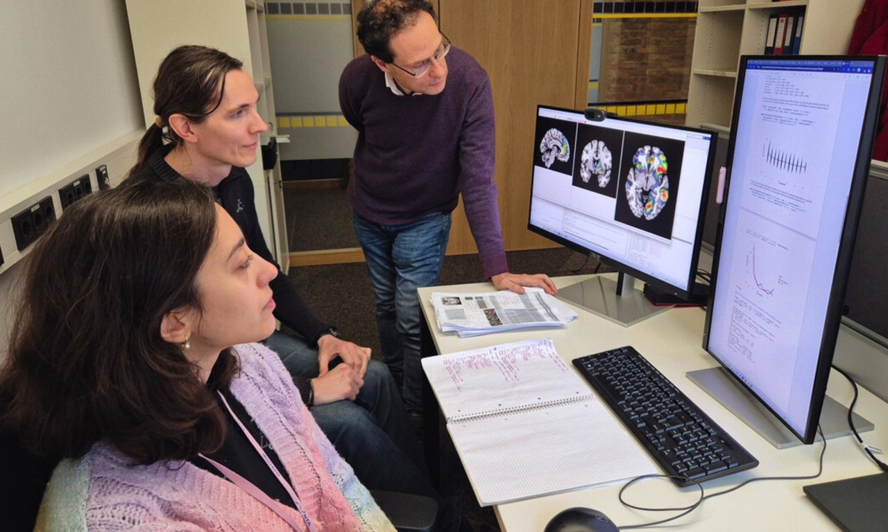 Three people stand or sit at a table and look at a screen.