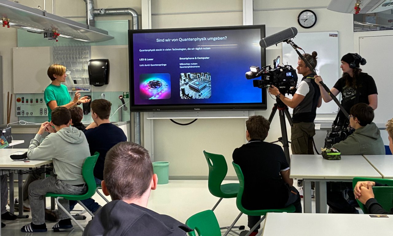 Klassenzimmer, Vortragende vor Whiteboard, im Vordergrund einige sitzende Schüler:innen