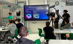 Klassenzimmer, Vortragende vor Whiteboard, im Vordergrund einige sitzende Schüler:innen