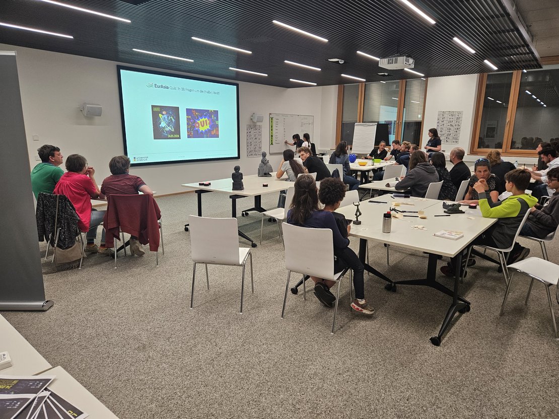 Seminarraum mit Menschen um Tische gruppiert, im Hintergrund sichtbar eine Leinwand mit der Quizankündigung und die Quizmasterin, die auf dem Whiteboard ein weiteres Team notiert