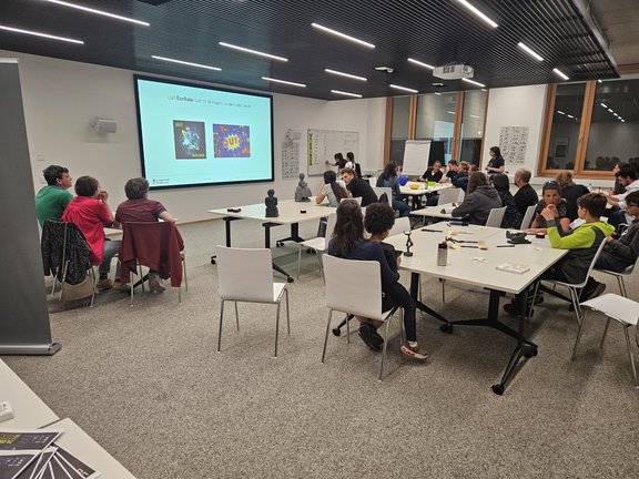 Seminarraum mit Menschen um Tische gruppiert, im Hintergrund sichtbar eine Leinwand mit der Quizankündigung und die Quizmasterin, die auf dem Whiteboard ein weiteres Team notiert