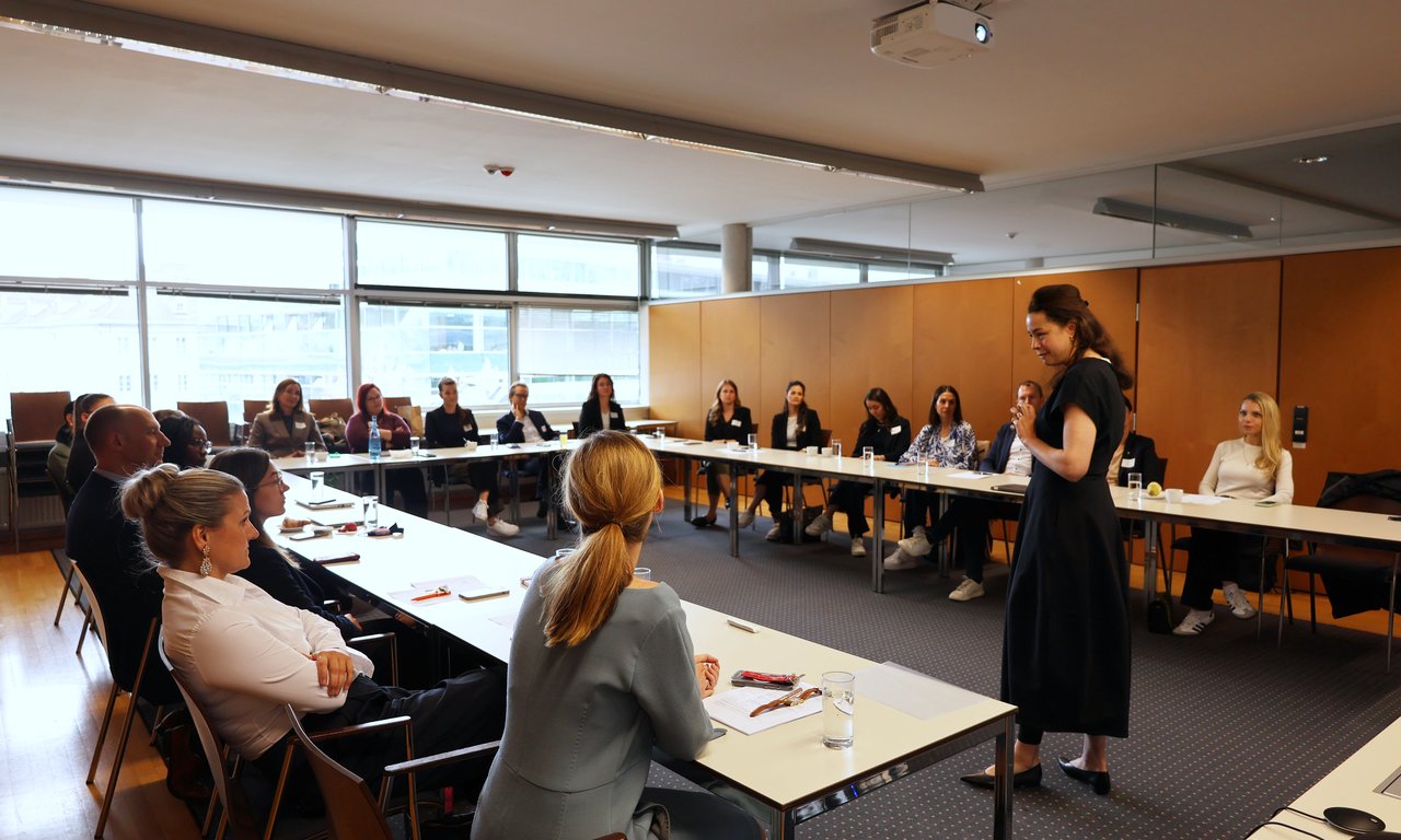 Mehrere Personen sitzen an Tischen in U-Form. Vorne steht eine vortragende Person.