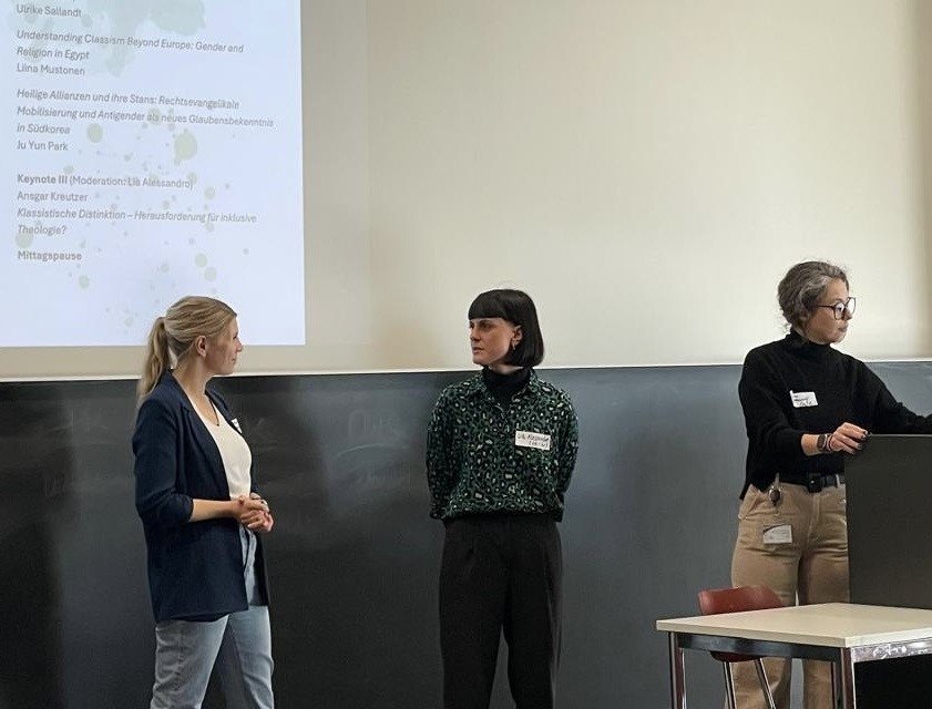 Zu sehen sind Carolin Hohmann (Münster/Innsbruck), Lia Alessandro (Frankfurt/Freiburg) und Jasmin Mausolf (Berlin). Sie stehen in einem Hörsaal, vor einer Tafel und halten einen Vortrag.