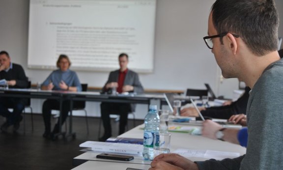 Personen in einem Besprechungsraum, im Hintergrund eine Präsentation auf einer Leinwand