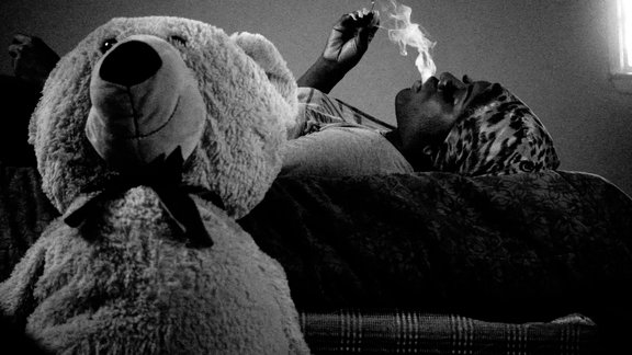SChwarz-weiß Bild einer Frau die auf dem Rücken liegend raucht. Auf der linken Hälfte des Bildes ist ein großer Teddybär zu sehen.