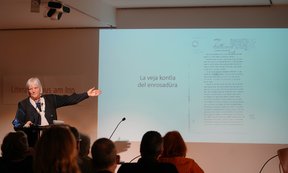 Ulrike Kindl steht an einem Rednerpult vor einer Leinwand und zeigt mit einer Hand nach oben.