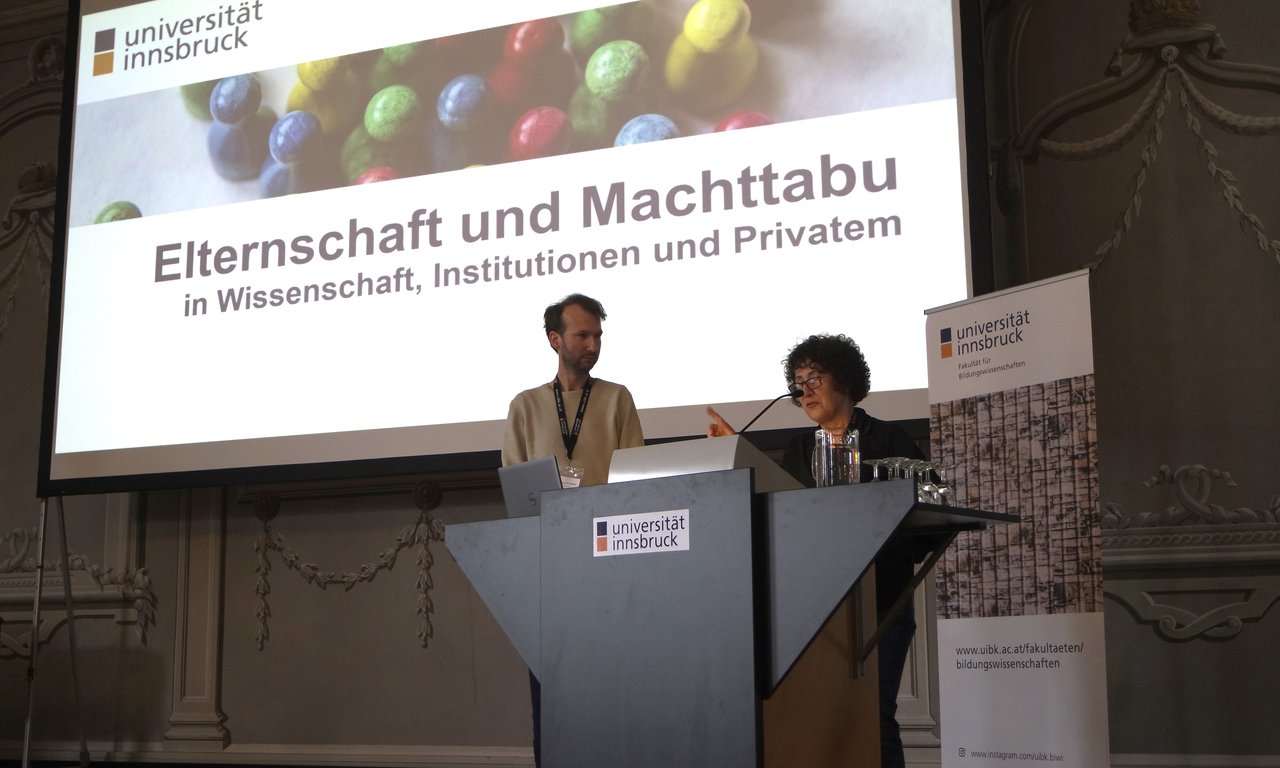 Zwei Personen hinter einem Rednerpult, hinter ihnen eine große Leinwand mit dem Namen der Tagung.