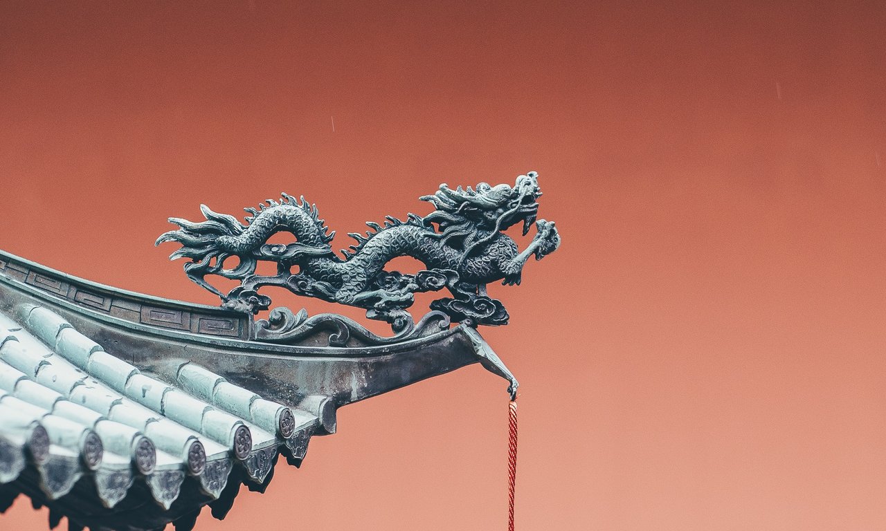 Ein Dachvorsprung mit einer Drachenskulptur und roten Bändern. Im Hintergrund eine rote Wand.