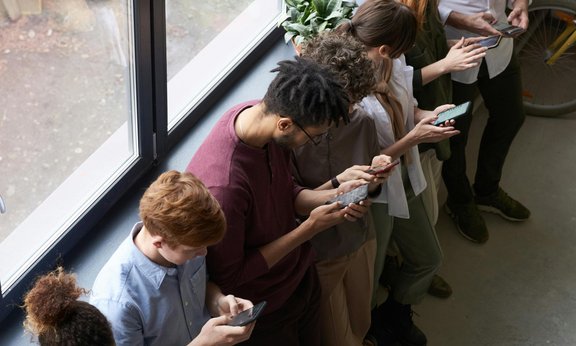 Mehrere Menschen lehnen an eine Wand, hinter ihnen Fenster, und schauen in ihre Handys.