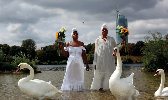 Zwei Frauen im weißen Kleid stehen in einem See umgeben von 3 weißen Schwänen