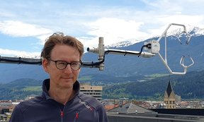 Thomas Karl stands in front of a measuring device, in the background a view over the city of Innsbruck with the Patscherkofel mountain
