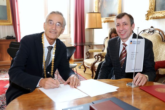 Zwei Männer sitzen an einem Tisch, einer unterschreibt ein Dokument