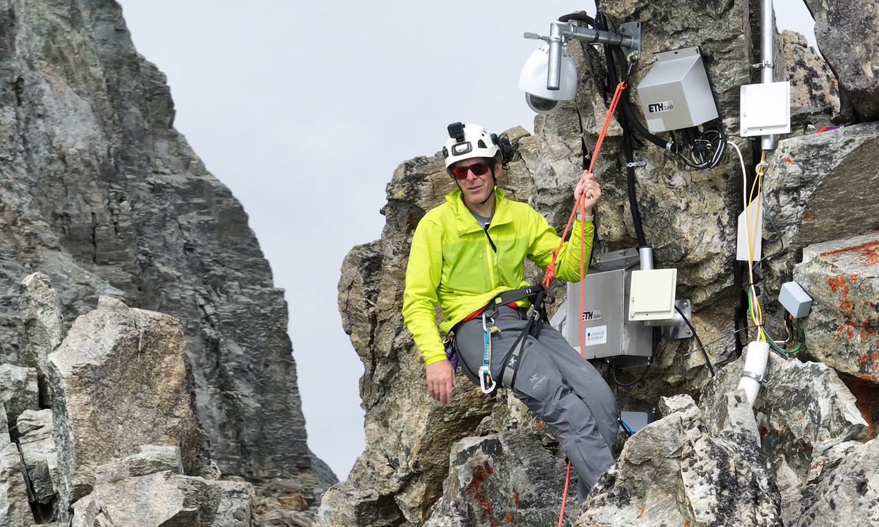 Ein Bergsteiger in gelber Funktionsjacke hängt an einem Seil im Fels, hinter ihm mehrere technische Geräte am Felsen.