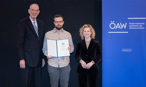 Drei Personen stehen nebeneinander, der mittlere von Ihnen hält eine Urkunde in der Hand