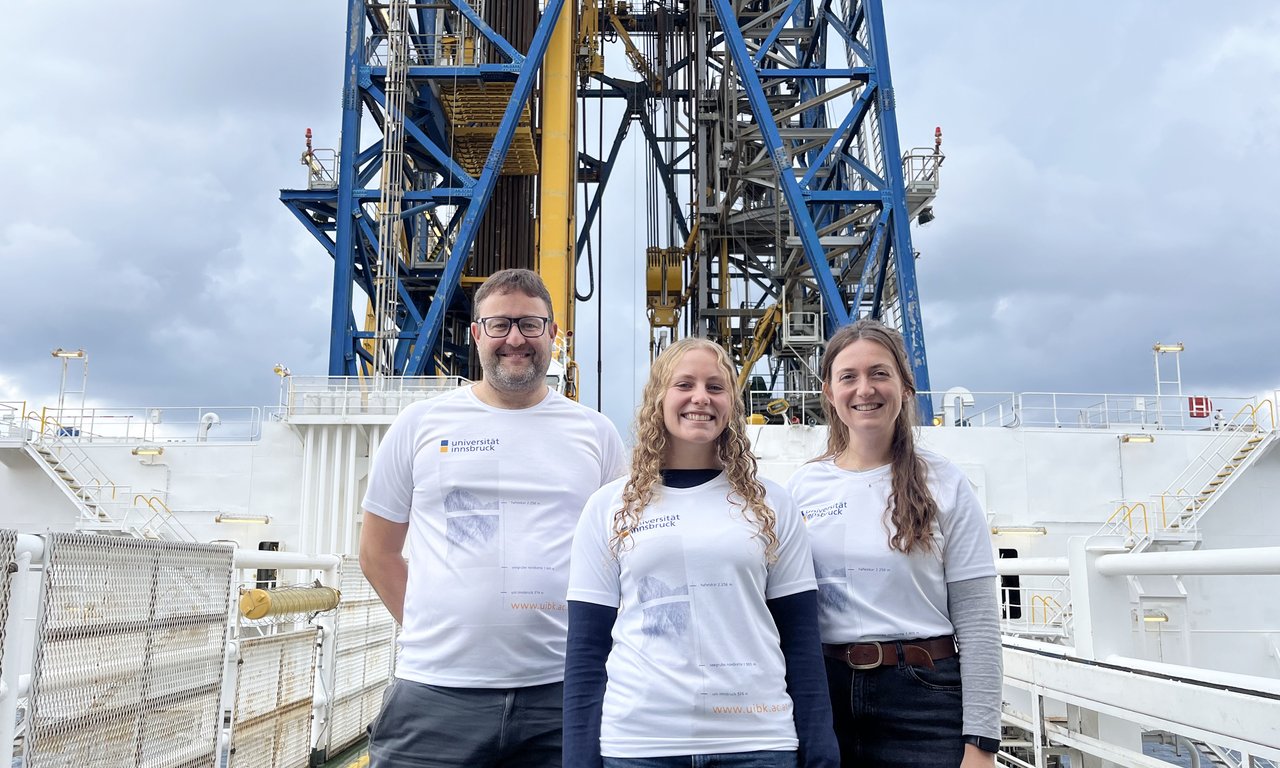 3 Personen in Uni-Innsbruck-T-Shirts stehen vor einem großen Bohrturm auf einem Schiff.