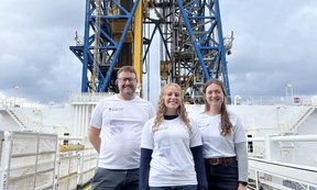 3 Personen in Uni-Innsbruck-T-Shirts stehen vor einem großen Bohrturm auf einem Schiff.