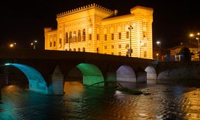 Die Vijećnica, das alte Rathaus von Sarajevo, ist ein Wahrzeichen der Stadt. Im Bild eine Ansicht des Gebäudes bei Nacht, fotografiert wurde vom Fluss Miljacka aus, eine Brücke im Vordergrund. Das Gebäude ist mit Scheinwerfern beleuchtet.