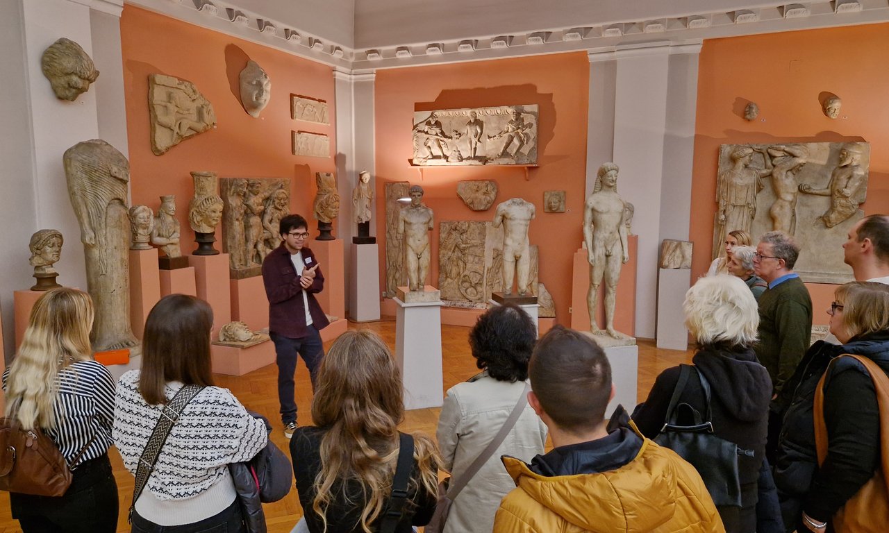 Mehrere Personen mit dem Rücken zur Kamera. Eine Person spricht zum Publikum. Im Hintergrund antike Statuen.
