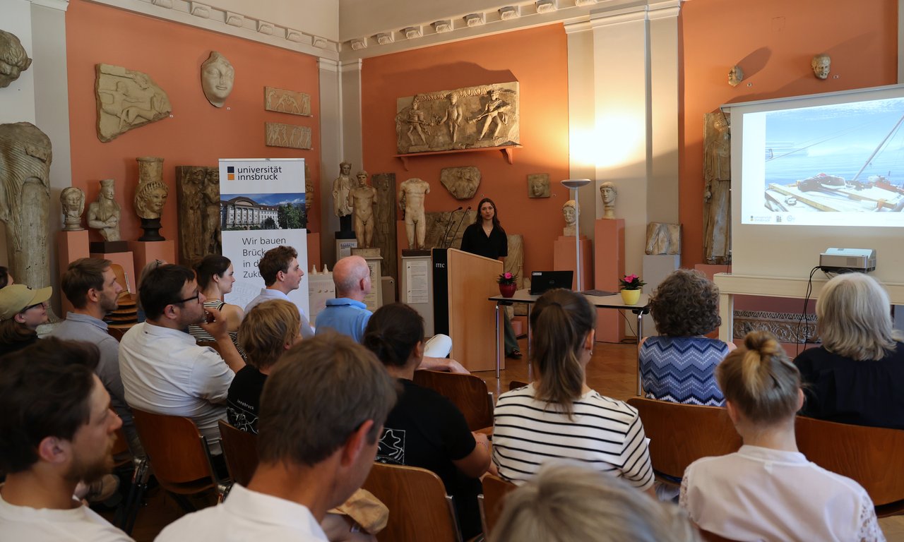 Publikum im Archäologischen Museum der Uni Innsbruck hört dem Vortrag einer Wissenschaftlerin zu.
