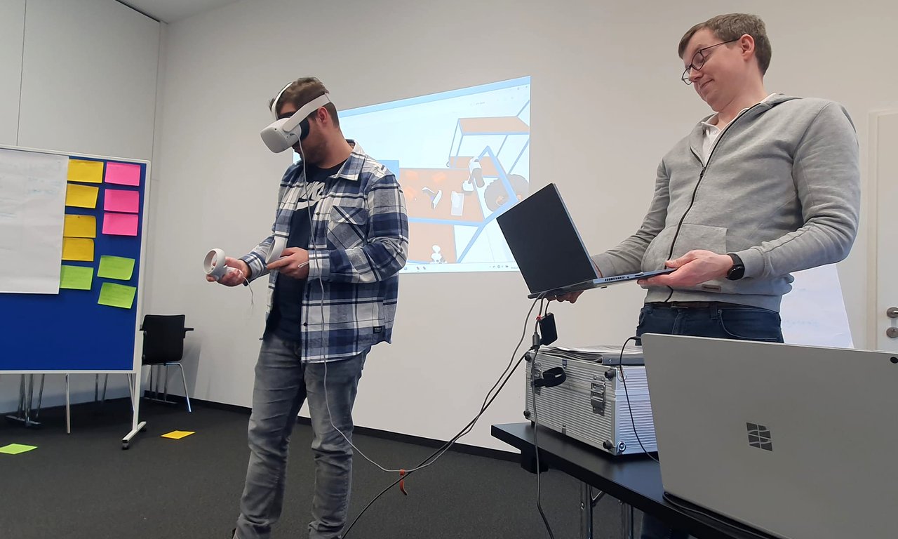 Mensch mit 3D-Brille im vorderen Bereich eines Seminarraums, ein zweiter Mann steht neben ihm, im Hintergrund ist projiziert, was er durch die Brille sieht.