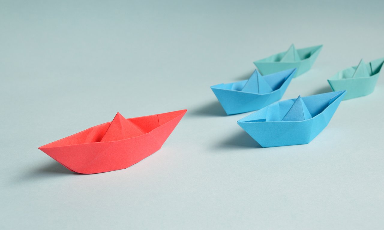 Symbolic image: Two blue and two green paper ships pursue a red paper ship on a white background.