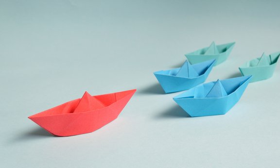 Symbolbild: Zwei blaue und zwei grüne Papierschiffe verfolgen ein rotes Papierschiff auf weißem Hintergrund.