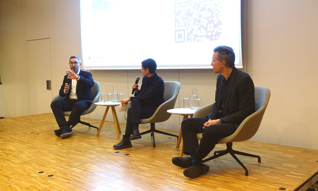 Drei Personen sitzen auf einem Podium und diskutieren.