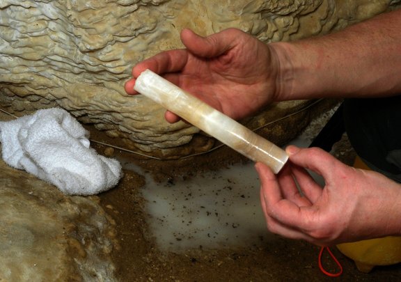 Two hands hold a long, narrow drill core made of shiny, semi-transparent rock with alternating white and brownish layers in a cave.