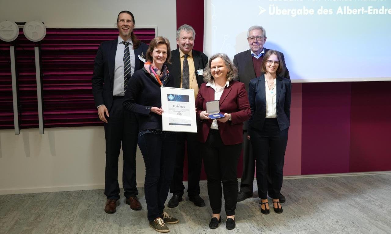 Gruppenfoto mit sechs Personen, zwei Frauen vorne halten eine Urkunde und eine Medaille.
