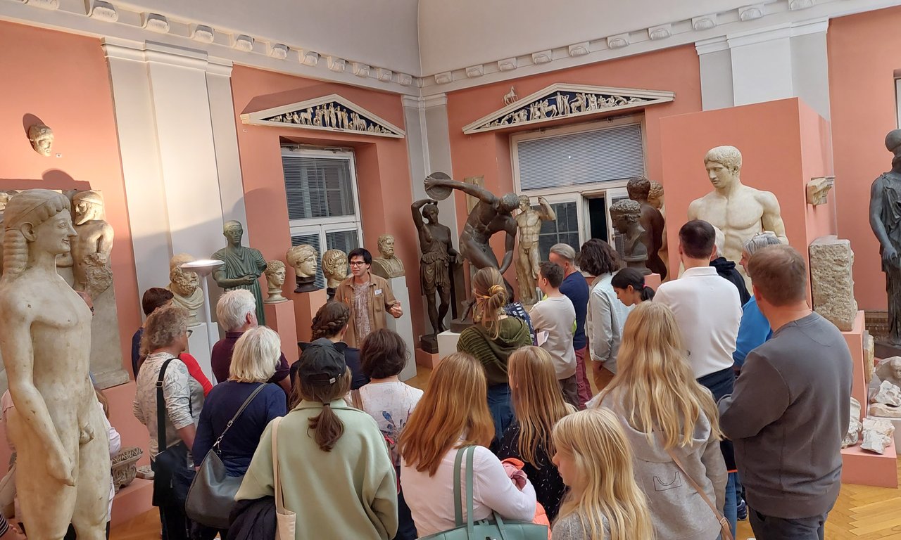 Viele Menschen stehen um eine männliche Person, die etwas erklärt. Um die Menschen herum befinden sich archäologische Statuen.