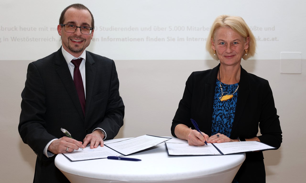 Zwei Personen stehen an einem Tisch und unterzeichnen Dokumente, beide blicken in die Kamera