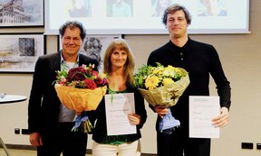 Drei Personen mit Blumensträußen und Urkunden blicken in die Kamera, dahinter eine Leinwand.