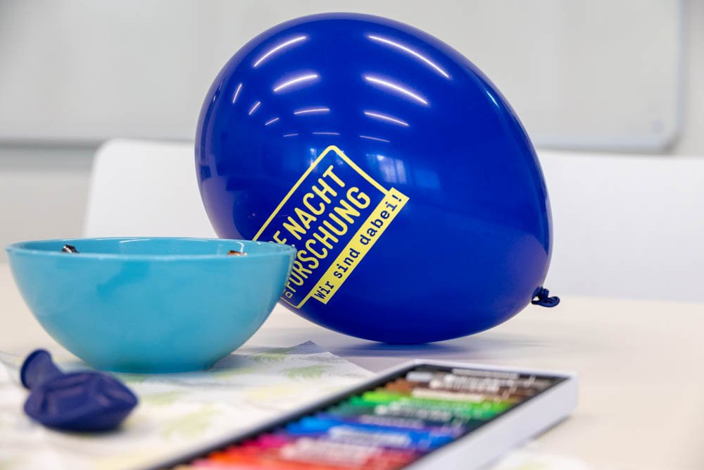 Wachsmalstifte liegen auf einem Tisch vor einem Ballon mit der Aufschrift lange Nacht der Forschung