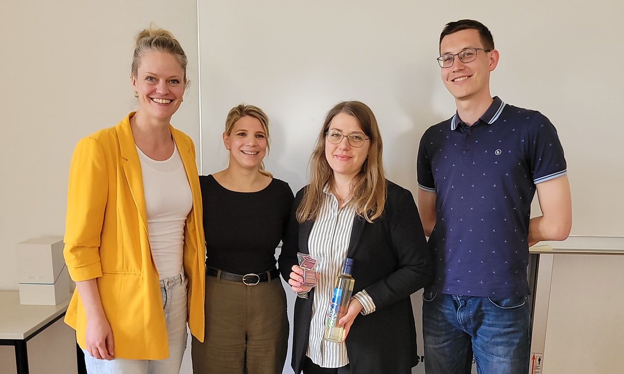 Zu sehen sind die Veranstalter_innen des Workshops. Sie stehen nebeneinander und lächeln. Eine der Personen hält ein kleines Geschenk in der Hand.