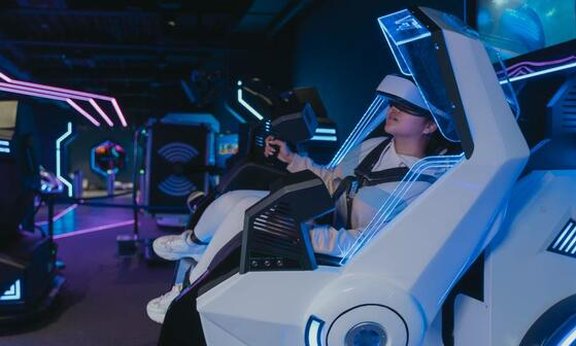 Mensch sitzt mit Spielekonsole und VR Brille in einem futuristischen Stuhl.