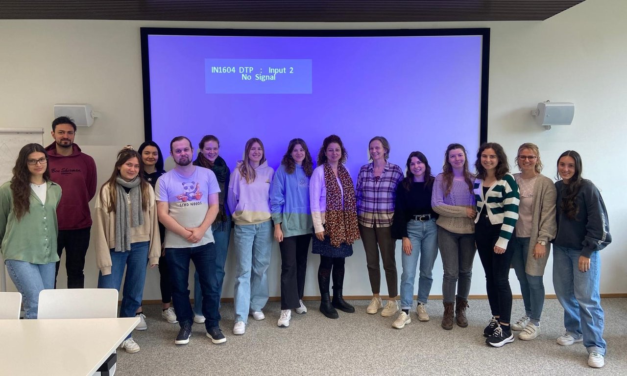 Eine Gruppe Studierender (15 Personen) steht in einem Seminarraum vor einer elektronischen Tafel.