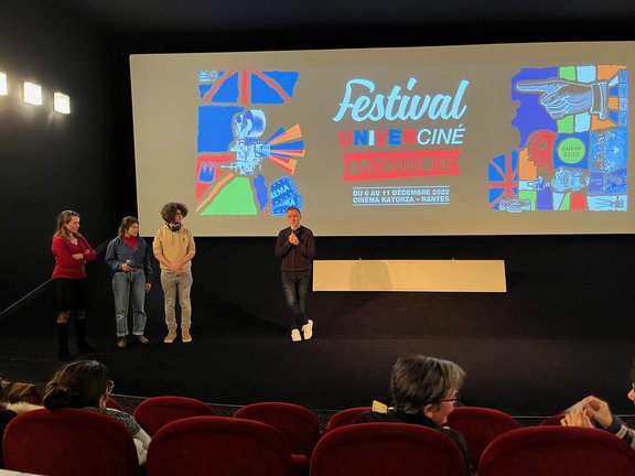 Ein Mann mit Mikro steht auf der Bühne eines Kinosaals, hinter ihm die Leinwand, drei weitere Personen stehen am linken Rand der Bühne.