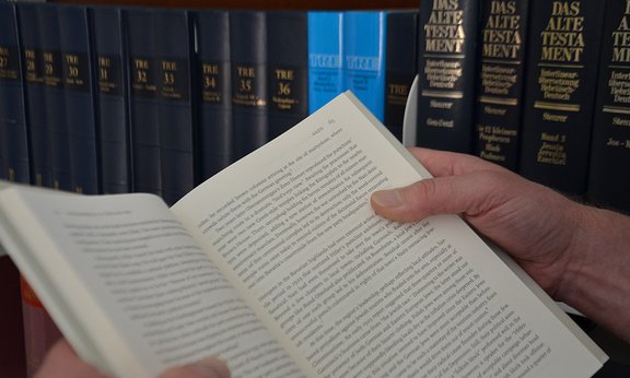 Jemand hält ein aufgeschlagenes Buch, im Hintergrund ein Bücherregal