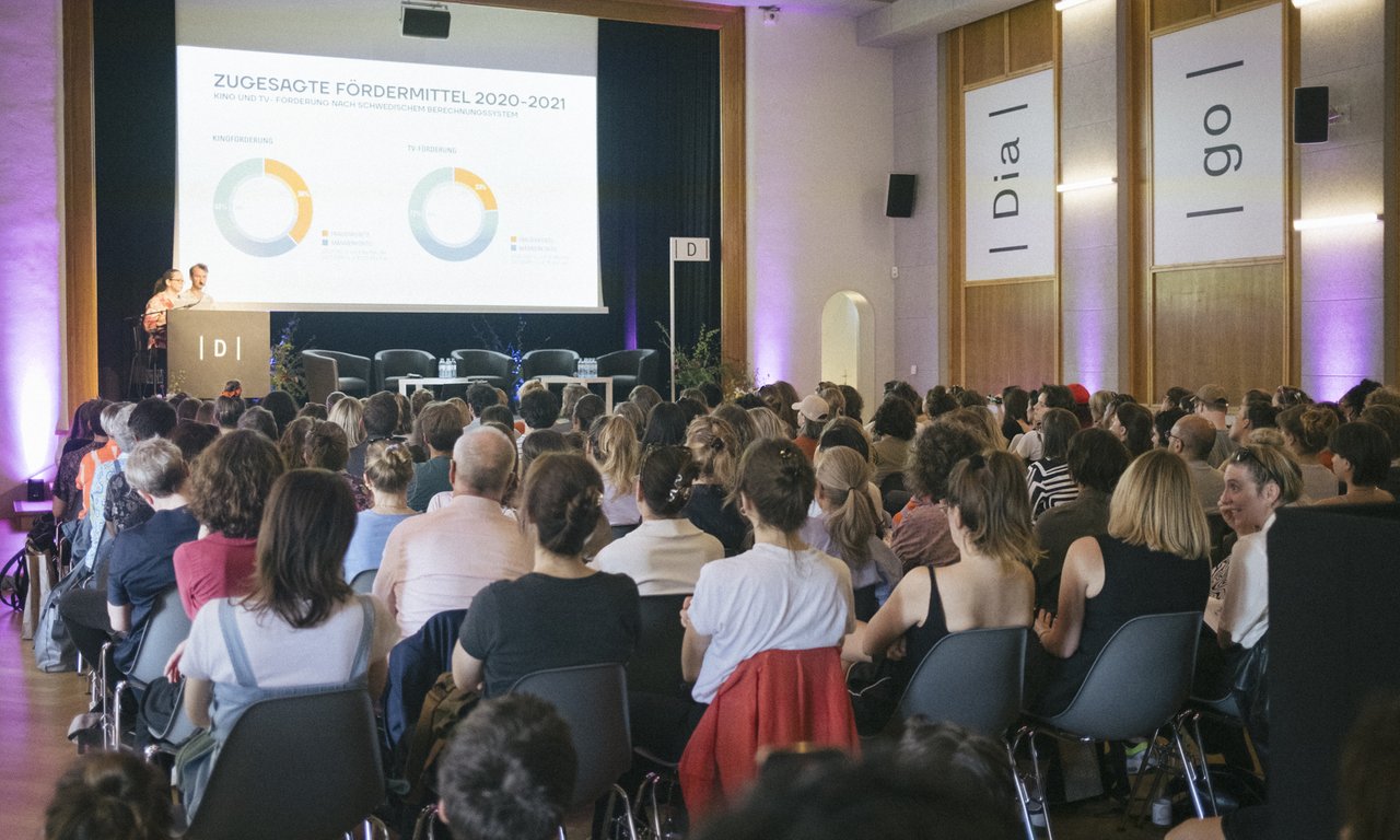 Voll besetzter Saal, Blick von hinten auf die Bühne. Man sieht die Hinterköpfe des Publikums, auf einer Bühne präsentieren zwei Personen.