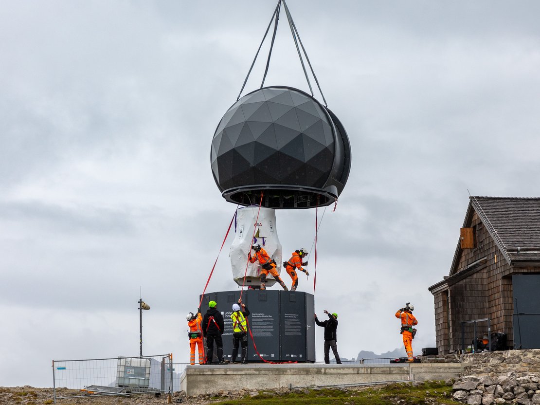 Eine schwarze Kuppel hängt an Gurten und schwebt dicht über dem Unterbau. Montagearbeiter ziehen die Kuppel in Position.